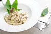 A white bowl of penne pasta with a creamy sauce and dark toppings, garnished with green leaves and small purple flowers, sits on a light wooden surface next to a cheese slicer. Produktbild-Vorschau