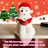 A Christmas-themed ghost figurine holding an open book, set against a festive background with Christmas trees and presents. Produktbild-Vorschau
