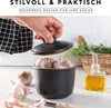 A hand is placing the lid on a black ceramic garlic keeper filled with garlic bulbs. Other garlic bulbs, a knife, and a bottle of olive oil are arranged around it, with a basket of rosemary in the background. Produktbild-Vorschau