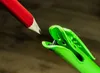 A close-up shot of a red pencil tip about to make contact with a black object held by a green plastic clip. Produktbild-Vorschau