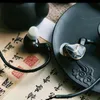 Two in-ear monitor headphones are placed on a surface with traditional East Asian calligraphy and a dark, decorative plate. Produktbild-Vorschau