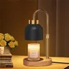 A candle warmer lamp with a black shade and gold accents sits on a wooden base, with a lit candle underneath. A bouquet of yellow roses is to the left, and stacked books are visible. Náhled obrázku produktu