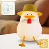 A cute, glowing duck-shaped night light with a hat sits on a white surface, with a blurred background of a couch and pillows. A small inset shows a close-up of the base of the lamp with regulatory markings. A timer icon is in the bottom right corner. Produktbild-Vorschau
