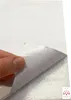 A hand is peeling back a corner of a white self-adhesive wall panel with a textured silver backing. Produktbild-Vorschau