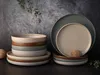A stack of ceramic bowls and plates in various earthy tones, arranged on a dark surface against a textured gray background. A sprig of greenery is visible in the upper left corner, and a small ball of twine rests on the plates on the right. Produktbild-Vorschau