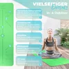 A woman is meditating on a green yoga mat outdoors on a wooden deck, with plants in the background. The mat has instructional markings. Produktbild-Vorschau