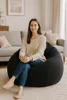 A woman sits on a black beanbag chair in a living room. Voir les images du produit