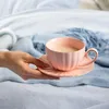 A hand holding a pink teacup and saucer on a bed with blue blankets. Produktbild-Vorschau