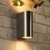 A close-up of a modern, cylindrical outdoor wall light mounted on a brick wall. Náhled obrázku produktu