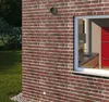 The exterior of a brick house with a window showing a kitchen interior. There is grass and foliage outside. Podgląd zdjęcia produktu
