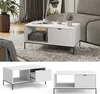 A white coffee table with a fluted drawer front is shown in a living room setting, next to a sofa and a plant. Voir les images du produit