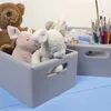 Several stuffed animals are in a gray wooden crate, with another crate and art supplies in the background. Produktbild-Vorschau