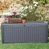 A dark gray storage box with a wicker-like texture sits on a green lawn, with potted plants on top and a bush with white flowers in the background. Produktbild-Vorschau