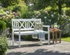 A white garden bench sits in a lush garden setting with flowers and greenery. Produktbild-Vorschau