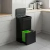 A black, multi-compartment trash can with a green inner bin, placed in a modern kitchen setting. Voir les images du produit