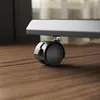 A close-up view of a black caster wheel attached to a gray metal frame, resting on a wooden floor. Anteprima dell’immagine prodotto