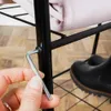 A hand is using an Allen wrench to tighten a screw on a black metal shelf unit. A pink folded fabric is on the shelf, and a black boot is visible in the background. Produktbild-Vorschau