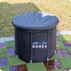 A black inflatable tub with a gray cover sits on a patio made of wood tiles and artificial grass. Produktbild-Vorschau