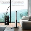A tall, white and black tower fan is positioned in a living room setting, near a sofa, with a large window in the background displaying a landscape. A remote control for the fan is also visible. Voir les images du produit