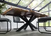 A live-edge wooden dining table with a black cross-shaped base, surrounded by chairs in a modern dining room with large windows. Produktbild-Vorschau