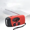 A red and black emergency hand-crank radio with an extended antenna and a flashlight on the front, set against a blurred background of mountains and sky. Podgląd zdjęcia produktu