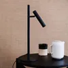 A black table lamp is placed on a black table next to a white teapot and a black and cream mug. The background is a textured light brown. Produktbild-Vorschau