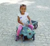 A young girl is riding a toy car with a trailer attached, outdoors on a cobblestone path. Produktbild-Vorschau