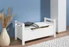 A white storage bench with a drawer and a lift-top lid is placed against a blue wall, next to a tall white pillar and a vase with dried grass. A patterned rug is visible on the floor. Produktbild-Vorschau