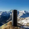 A black thermos bottle sits on a rock with snow-capped mountains in the background. Náhled obrázku produktu
