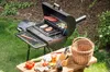 A barbecue grill with food cooking on it, set up outdoors on a wooden table with various ingredients and a basket. Podgląd zdjęcia produktu