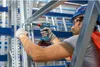 A person wearing a hard hat and safety glasses is using a cordless impact wrench to assemble a metal shelving unit in a warehouse. Produktbild-Vorschau