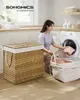 A woman is loading laundry into a washing machine in a laundry room. A large woven laundry basket is in the foreground. Produktbild-Vorschau