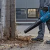 A person is using a leaf blower to clear leaves from the ground next to a tree. Produktbild-Vorschau