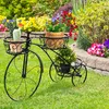 A black metal bicycle-shaped plant stand with three baskets holding potted plants, set in a garden with colorful flowers in the background. Produktbild-Vorschau