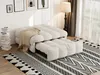 A beige sofa bed is shown in a living room setting, partially pulled out into a bed, with a blanket draped over it. A patterned rug is on the floor, and a stack of books is to the left. A floor lamp and curtains are visible in the background. Produktbild-Vorschau