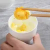 A hand holding chopsticks is lifting a piece of cooked egg from a white cup. Produktbild-Vorschau