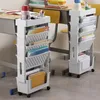 Two white rolling storage carts filled with books and folders are placed next to school desks. Produktbild-Vorschau