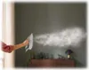 A hand holding a steam iron, emitting steam towards a wall in a room. Furniture and a lamp are visible in the background. Voir les images du produit