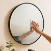 A hand is wiping condensation off a large round mirror with a white cloth. Produktbild-Vorschau