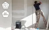 A construction worker is on a ladder installing drywall, with a portable heater in the foreground. Produktbild-Vorschau