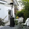 A patio heater stands next to a small table and chairs on a paved patio, with lush greenery in the background. Produktbild-Vorschau