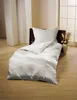 A white duvet and pillow set laid out on a wooden floor, with decorative pillars and a window in the background. Náhľad obrázku produktu