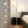 A floor lamp with two glass globes is shown next to a nightstand and a bed. Náhľad obrázku produktu