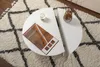 A white, semi-circular coffee table is placed on a fluffy rug with a geometric pattern. A small candle and a magazine are on one half of the table, and a vase with dried branches is on the other half. Produktbild-Vorschau