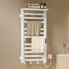 A white electric towel warmer mounted on a light brown wall, with two towels draped over it. There are also some bottles and rolled towels on a shelf above the warmer, and a small vase with dried flowers on a cabinet to the right. Anteprima dell’immagine prodotto