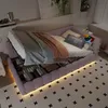 A pink upholstered bed with storage drawers, featuring a mattress, blankets, and pillows, set in a bedroom with a patterned rug and wall art. Produktbild-Vorschau