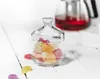 A small glass jar filled with colorful gummy candies sits on a white wooden surface. A teapot with red liquid is visible in the background. Náhled obrázku produktu