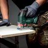 A person wearing work gloves is using a green and black Metabo cordless jigsaw to cut a piece of white laminate-covered particle board resting on a table. Podgląd zdjęcia produktu