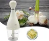A white garlic chopper is shown in the foreground, with fresh garlic bulbs and herbs in the background. Náhľad obrázku produktu