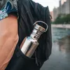 A person's arm and shoulder are visible, with a black backpack on their back. A stainless steel water bottle with a bamboo lid is tucked into a side pocket of the backpack. The background shows a blurred cityscape and water. Podgląd zdjęcia produktu